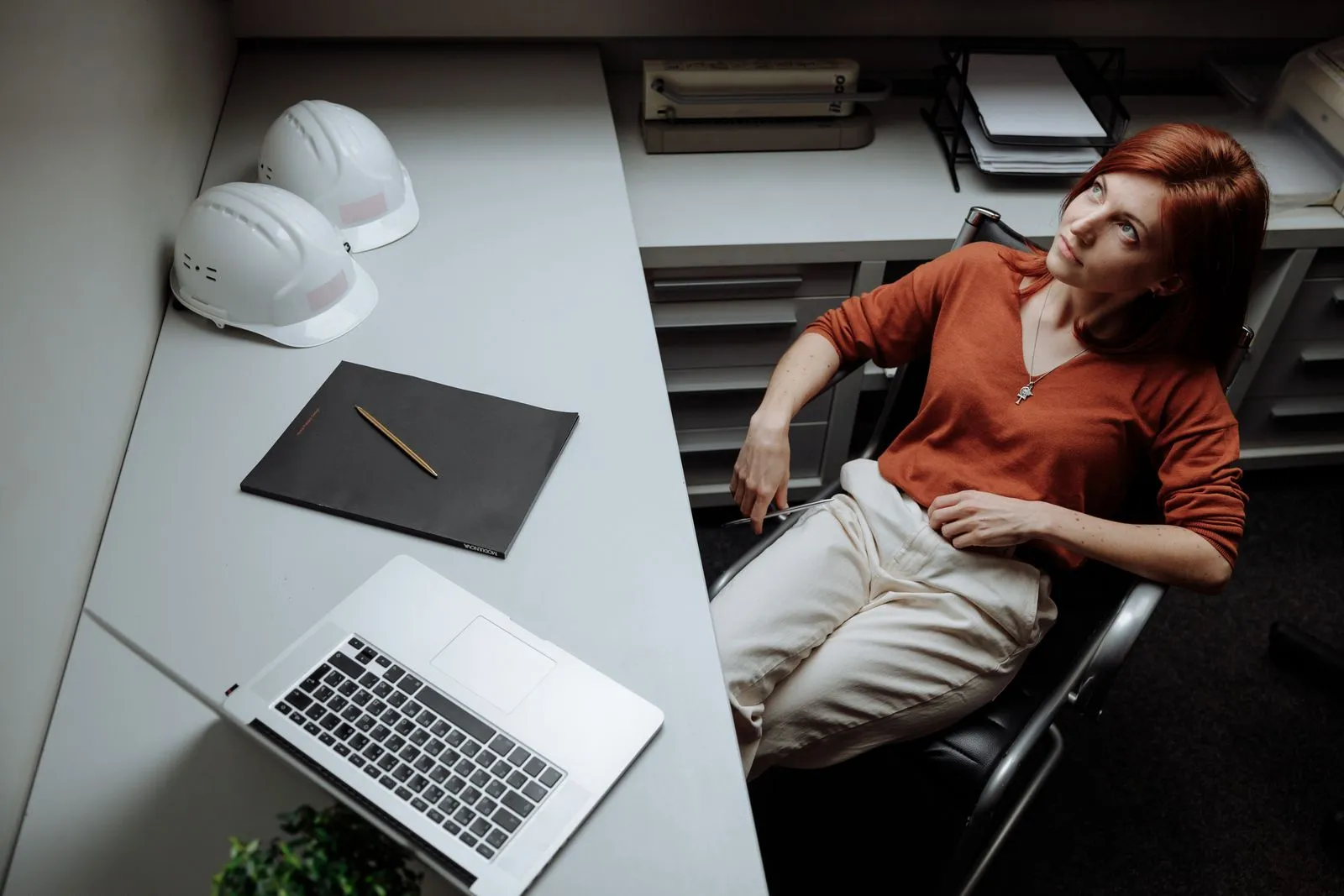 Professional woman relaxing at desk with laptop, hard hats, and documents, contemplating ideas.